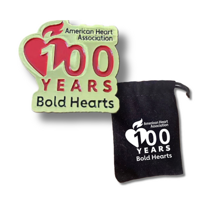 lapel pin of AHA Bold Hearts 100 Years logo, gold with red and black recessed design next to black velvet-like bag with logo in white